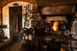 Interior of a blacksmith forge. There is a fire roaring in the furnace and a wide variety of forging tools hanging on the wall