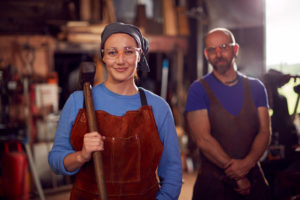 two blacksmiths, one male and one female standing in a forge. The female is slightly in front of the male, wearing a brown protective apron, and holding a forging hammer in her right hand.